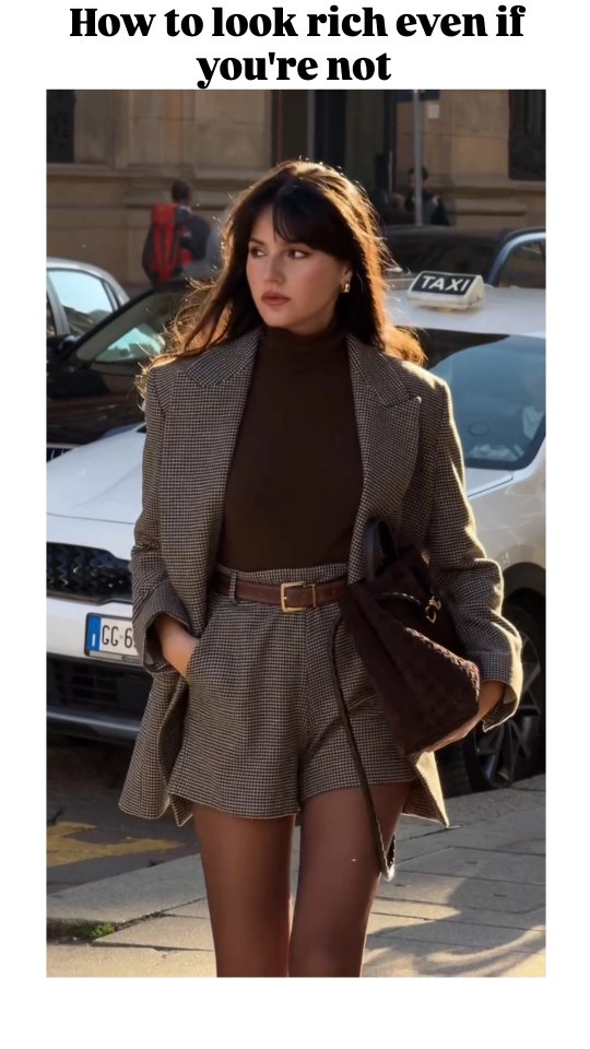 A woman in a stylish outfit is walking down a city street. She is wearing a patterned blazer and shorts with a brown turtleneck and carrying a dark purse. The background includes parked cars and buildings.