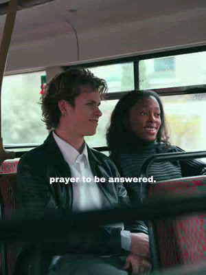 A medium shot of two people sitting on a bus. A young man with fair skin and short, wavy hair is on the left. He is wearing a black jacket over a white shirt, looking off to the side with a slight smile. Next to him is a dark-skinned woman with her hair pulled back. She is wearing a black turtleneck sweater and smiling. Below them is a red and black striped bus seat.