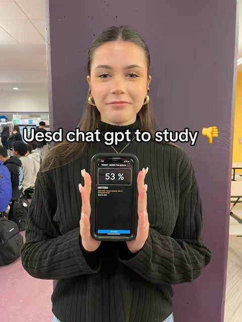 A young woman with long hair and a black sweater stands in a hallway, holding a smartphone displaying an app's interface. The background includes a purple pillar, providing context.