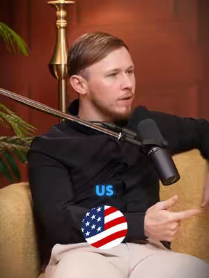 A man with short, light brown hair and a black shirt is seated on a tan-cushioned surface. He is holding a microphone and pointing to the right. The image includes a US flag emblem and the word "US". Behind him, a gold-colored object resembling a lamp base is visible, and a plant with green leaves adds a touch of nature to the background.