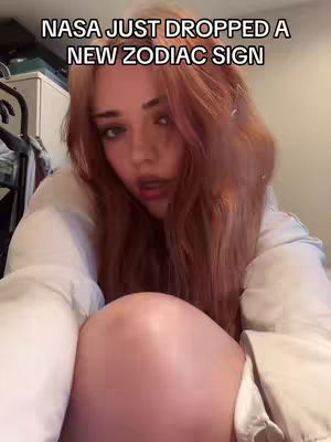 A young woman with long, reddish-brown hair is the central focus, captured in an intimate, close-up shot. She's leaning towards the camera, with her arms wrapped around her own knees, giving the shot a sense of closeness. The background is soft-focused, suggesting a bedroom setting, which creates a warm and personal atmosphere. The lighting is soft and diffused.