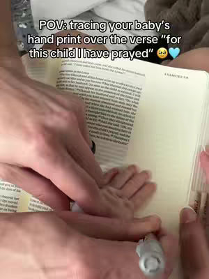 A person's hand traces a baby's hand print over a page of a book. The text on the book appears to be scripture. The baby's tiny hand is visible beneath the adult hand, with the pen tip pointing to the page. The lighting is soft and natural.