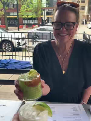 A woman with glasses and sunglasses on her head smiles while holding a cocktail with a lime. The setting appears to be an outdoor restaurant.