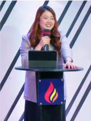 A woman with long brown hair stands behind a black podium, holding a microphone and smiling. The background features a white and black geometric pattern. The podium has a logo depicting a flame.