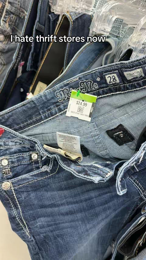 The image shows a close-up of used jeans hanging on a rack. The focus is on a pair of blue denim jeans, with visible wear and tear. A price tag is clipped to the jeans, indicating they are for sale. The overall composition highlights the texture and condition of the used clothing item.