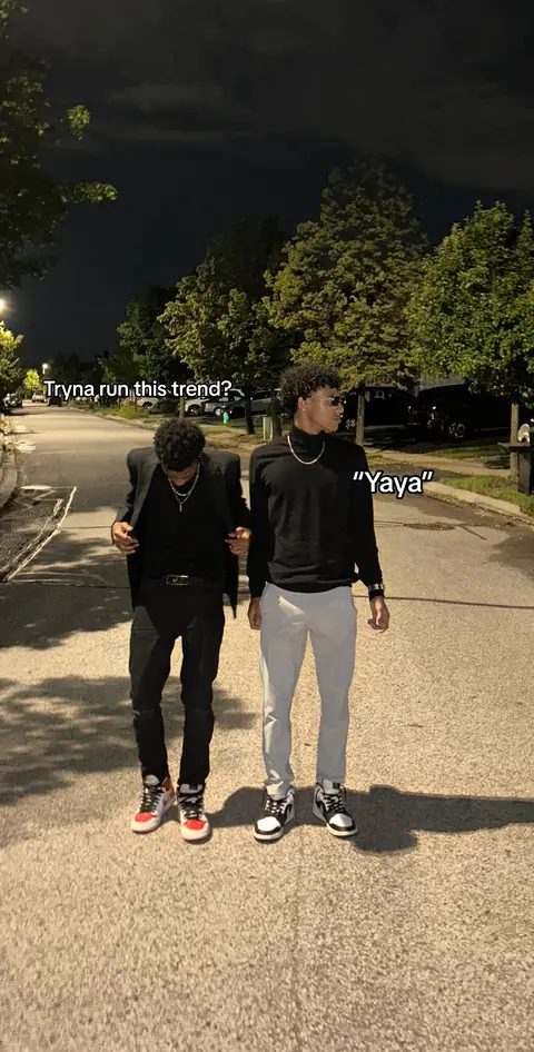 Two young men stand side-by-side on a paved pathway at night, appearing to walk towards the viewer. The man on the left wears a black blazer over a black top and black pants, paired with red and white sneakers. The man on the right is dressed in a black turtleneck with light gray pants, and sneakers. Text overlays the image, and the background shows trees and dimmed streetlights.