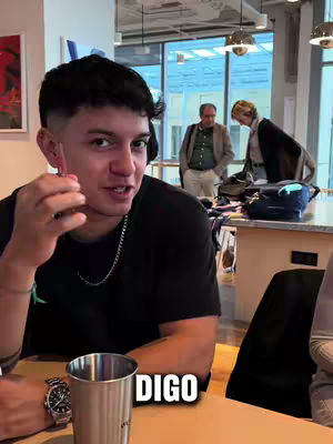 A young man with short dark hair is smiling directly at the camera. He's wearing a black t-shirt and a silver chain necklace and the text "DIGO" in a bold black font with a white outline is near the bottom of the screen. In the background are blurred figures of people in an office or a similar setting.