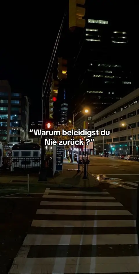 A nighttime city scene. A pedestrian crosswalk, marked with white stripes, leads towards the center of the frame. Buildings and streetlights illuminate the background. Text overlayed on the image.