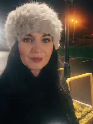A woman in a fur hat is the focus, seen from the chest up. The background is blurred, with streetlights and a muted cityscape visible. The color palette is cool, with blacks and whites dominating; the hat is a light gray, and her skin has a natural tone with touches of makeup. The composition centers on her face, conveying a mood of winter and a casual, outdoor setting at night.