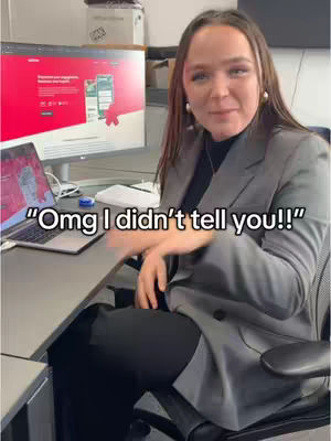 A woman at a desk is smiling at the camera. Behind her is a computer screen and a laptop on the desk. Text overlayed states, "Omd I didn't tell you!!"