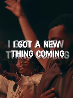 A medium shot captures a group of people in a darkened environment, likely a concert or church service, with arms raised. The focus is on the text overlayed on the image, which is a blurred white semi-transparent text that says: I GOT A NEW THING COMING. The lighting is warm and focused on the figures, creating shadows and a sense of depth.