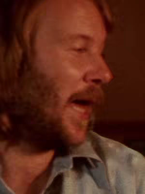 A close-up shot of a man with light skin, looking towards the left with his mouth open, suggesting he is speaking. He has a beard and light-colored hair. The background is dark, with warm brown tones.