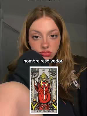 A young woman with long brown hair is centered in the frame, looking down slightly with a neutral expression. She's wearing a dark hoodie. In front of her is a tarot card, featuring a person in red robes and a gold crown. The background is a plain, neutral color, which makes the subject and the card the focus.