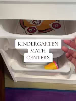 The cover image shows a close-up view of a white storage compartment or drawer, which is partially open, revealing various items inside. At the bottom of the frame, there is a colorful toy. In the middle drawer, there are several cards with dot patterns and the number 6. The upper portion is obscured, while the lower section shows a small, yellow and pink toy. Text overlayed on the image says 'KINDERGARTEN MATH CENTERS'. The background exhibits a gradient of purple and potentially other colors like green on the bottom as well.