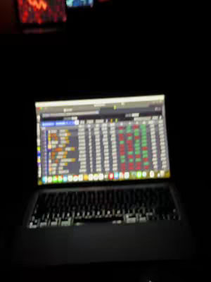 A laptop is open on a desk in a dimly lit area. The screen displays a trading platform interface with charts and numerical data, suggesting financial analysis or trading activity. The laptop keyboard is visible in the foreground, and the overall composition focuses the viewer's attention on the trading platform.