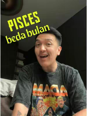 A selfie-style video with the creator, a man with short black hair and an open mouth expression. He's wearing a dark t-shirt with a multi-colored graphic. Above him, the words \"PISCES\" and \"beda bulan\" are written in the image in a yellow, stylized font. The background is a dark wall.