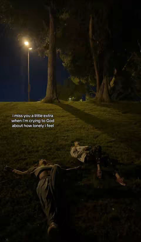 Two people are lying on a grassy hill at night. The scene is dimly lit, with a street light casting a soft glow. Text overlay reads: "i miss you a little extra when i'm crying to God about how lonely i feel."
