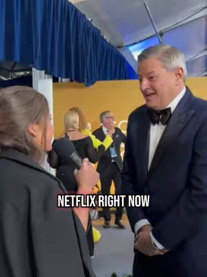 A woman with dark hair and a black top holds a microphone in front of a man in a suit with a bow tie. The man is smiling. The background features a yellow wall and people. Text that reads NETFLIX RIGHT NOW is present.