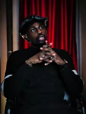 A man with a dark complexion is seated in front of a red curtain. He's wearing a black hat and sweater, with his hands clasped in front of him. His mouth is open as if he is speaking, capturing a moment of apparent dialogue or deliberation.