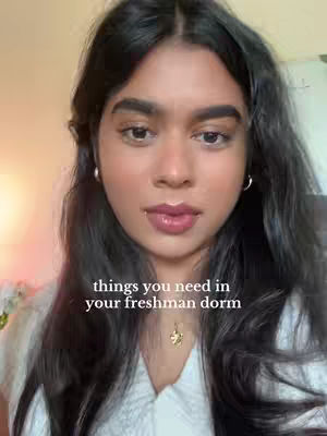 A close-up shot of a young woman with long, dark hair and fair skin. She is looking towards the camera. The text overlay reads "things you need in your freshman dorm."