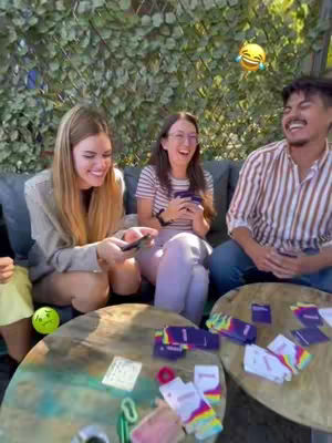 Three friends smiling and laughing while playing a card game outdoors. They are sitting around a wooden table with game cards and accessories spread out. A green backdrop decorated with leaves and a sky with sunshine in the background.