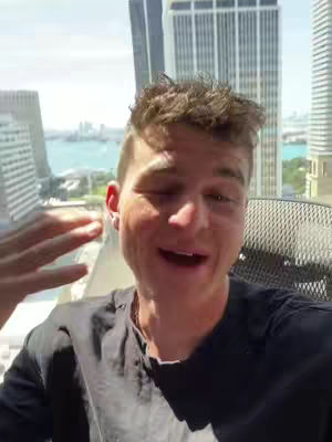 A man is smiling, his face and upper body filling the frame. He's in front of a large window with tall buildings and water visible in the background. The color palette is bright, reflecting daylight.