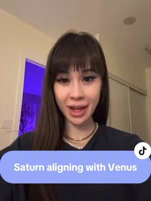 A young woman with long brown hair and bangs is centered in the frame, looking directly at the camera. She wears a black top and a necklace. A blue bar with white text that reads "Saturn aligning with Venus" is overlaid at the bottom of the image.