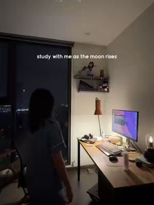 A person stands in a room with a large window looking out at a city at night. The room has a desk with a computer and a lamp.