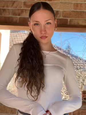 A young woman with long, dark curly hair gathered in a low ponytail, stands in front of a brick wall and a window. She wears a white long-sleeved top. Her gaze is directly at the camera.