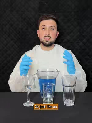 A man in a white lab coat and blue gloves stands in front of a black background. Various beakers and glassware are on a table in front of him, with a large beaker containing blue liquid.