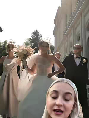The cover image showcases a wedding scene. In the foreground, a young woman with blonde hair is positioned with her head near the bottom of the frame, looking at the camera and speaking. In the middle ground, the bride, in a white wedding dress and veil, holds a bouquet and is flanked by bridesmaids. Groomsmen and other guests are in the background. The setting appears to be an outdoor area, possibly near a building, with sunlight creating a bright and airy scene.