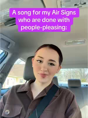 A woman is seated in a car, looking towards the camera with a light smile. She's wearing a brown collared shirt and a seatbelt. Above her head is a rectangular purple banner with white text on it.