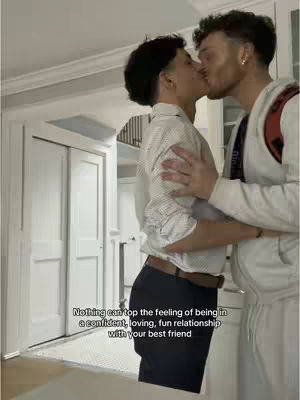 Two men are embracing and kissing. They are in an indoor setting with white walls and doors visible in the background. One man wears a white shirt and black pants, while the other has a white jacket with a red backpack strap. The overall composition is warm and intimate.