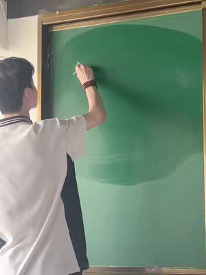 A person is writing on a green chalkboard, viewed from behind. The person is wearing a white shirt with black trim and has a brown watch on their wrist. The background is a simple wall.