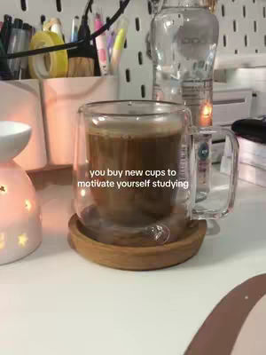 A close-up shot features a clear glass mug filled with coffee, resting on a small wooden coaster. The mug and coaster sit on a white surface with a blurred background of a study desk, including stationery and a water bottle. Text overlayed reads: 'you buy new cups to motivate yourself studying'. The composition is well-lit with warm tones.