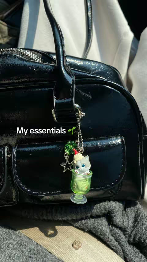 A close-up shot of a black, glossy handbag with a silver zipper on top. Dangling from the handle is a charm featuring a cat figurine inside a green glass. The text 'My essentials' is overlaid near the bottom left.