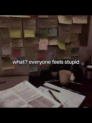 A dimly lit workspace or study area. A corkboard covered with various colored sticky notes dominates the background. In the foreground, open papers and pens are laid out on a dark wooden surface, with a coffee mug present to the right. The overall tone is focused yet relatable.