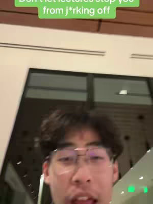 A selfie-style shot featuring a young man with short, dark hair and glasses. He is looking directly at the camera and talking. Above him is a green text overlay. The background is blurred, showing an indoor environment with neutral colors.