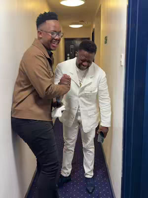 Two men, one in a brown jacket and the other in a white suit, are in a hallway. They are smiling and shaking hands, creating the impression of a friendly interaction.