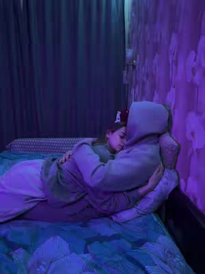 A dimly lit image features two people embracing in bed. One person is wearing a gray hoodie with the hood up, and the other is nestled against them. The scene is bathed in soft, cool-tone lighting, with purple hues dominating the background and adding to the intimate setting. The bed has a blue patterned bedspread, and there is a curtain in the background. The overall composition is calm and comforting.