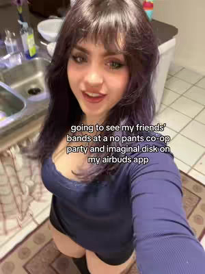 A woman is shown in a selfie-style shot, angled from above. She is indoors, with a sink, countertop, and other kitchen items in the background. The woman has dark hair, is wearing a blue top, and appears to be speaking to the camera. Overlay text adds context to the scene.