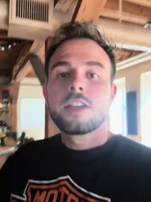 A medium shot shows a man's face. He is looking directly at the camera, with a casual expression. The background includes visible wooden beams and architectural details suggesting an indoor setting, possibly a home or a restaurant. The man is wearing a black t-shirt.