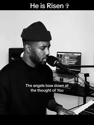 A grayscale image features a Black man playing a piano and singing into a microphone. The text overlay "He is Risen" appears at the top with a cross symbol. Additional text "The angels bow down at the thought of You" is also visible.