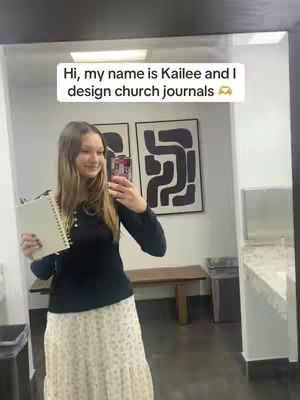 A young woman with long hair stands in front of a mirror, holding a notebook and a phone. She is smiling. Behind her are abstract artworks and bathroom fixtures.
