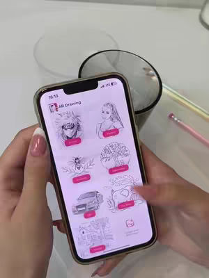 A person holds a smartphone displaying the 'AR Drawing' app interface. The phone, with a gold-colored frame, is positioned in the foreground, slightly angled towards the viewer. The app interface features various drawing options with colorful icons. The scene is set on a white surface, with a transparent container and a pencil visible in the background. The person has manicured nails.