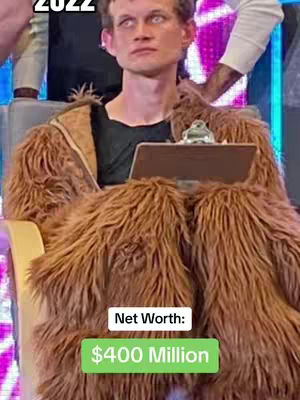A man wearing a brown furry costume, seated in a chair behind a table. Two text boxes are overlaid indicating net worth of 400 million USD.