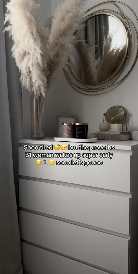 The cover image shows an interior scene with a neutral color palette. A white dresser with four drawers is the central element, with two decorative candles set on top of it. A vase with feathery pampas grass, a round gold mirror, and a stack of decorative books. The text overlayed at the top reads "Sooo tired 😴 but the proverbs 31 woman wakes up super early 😴😂🏃♀️🥺 sooo let's goooo."