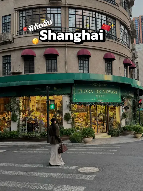 A woman is walking across a crosswalk in front of a building and vibrant shop. The words "Shanghai" are prominently displayed on the image with a couple of emoji and a flag.