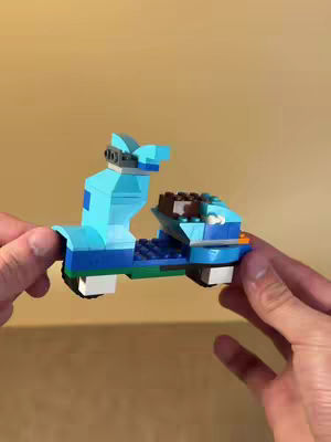 A person holds a small LEGO model of a scooter against a plain beige background. The scooter is mainly light blue with darker blue and some green and brown accents. The person's hands are visible, holding the model on either side.