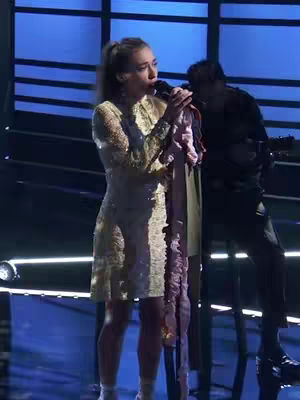 A woman with a ponytail stands on a stage, singing into a microphone. A person plays guitar in the background. The stage is lit with blue lights.